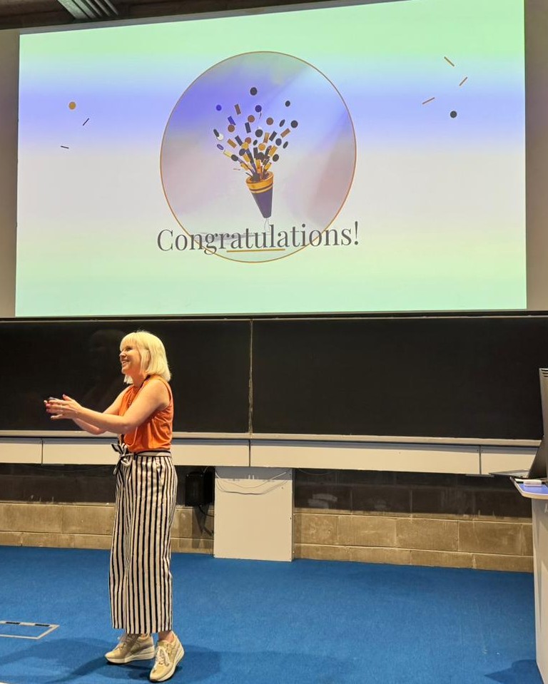 Brooke Gallagher smiling at the front of a Trinity College Dublin lecture theatre as a projected slide reading ‘Congratulations!’ closes the session behind her.